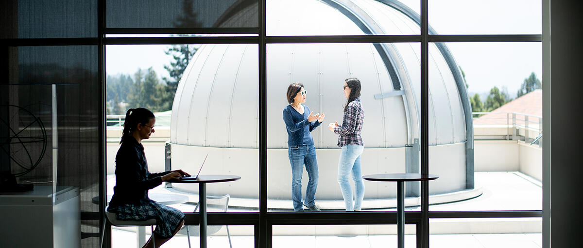 Photo of Chung-Pei Ma and student through window in front of a telescope dome, while another student sits working at a table inside