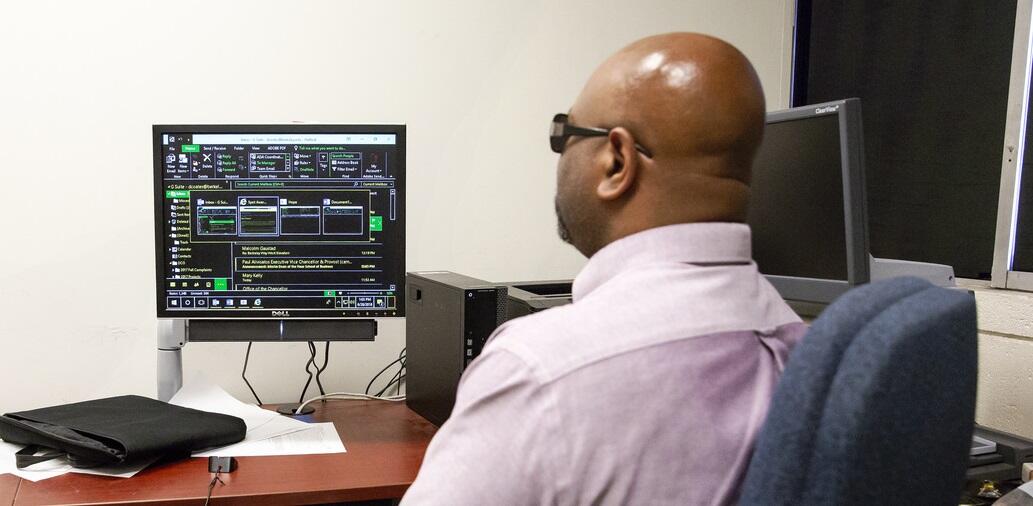 Computing Resources Man in pink shirt and dark eyeglasses, seated at desk, looking at computer monitor