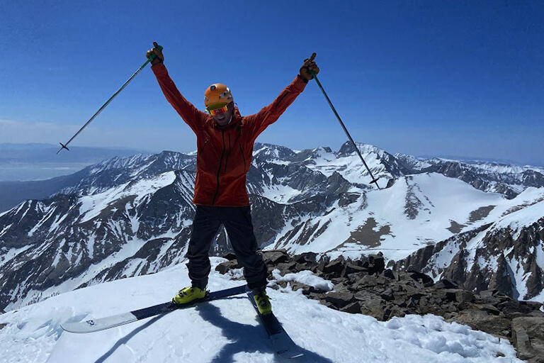 Roger Romani waving on skis at the top of a snowy summit